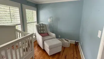 Small nursery corner with a white glider chair, footstool, lamp, and basket by a blue wall and windows.