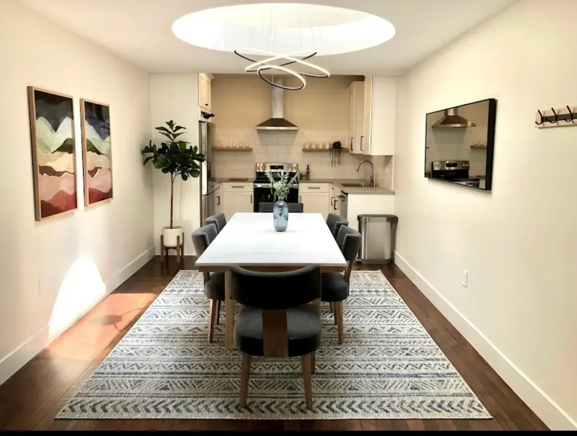 Modern dining room with a long table, patterned rug, wall art, and a small kitchen in the background