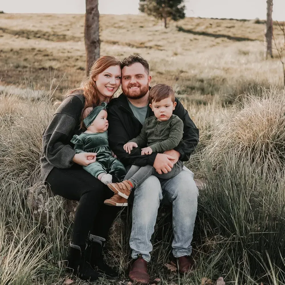 Family of four sitting together outdoors in a grassy field, smiling at the camera.