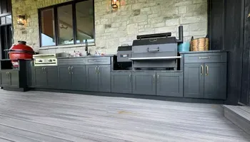 Outdoor patio kitchen with dark cabinets, grill, and red smoker against a stone wall