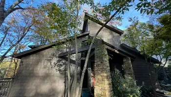 Weathered wooden house with a steep roof and stone chimney, surrounded by trees in daylight