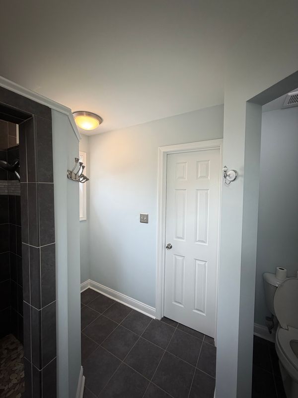 Small bathroom with dark tiled floor, white walls, vanity light, closed door, and partial shower and toilet visible.