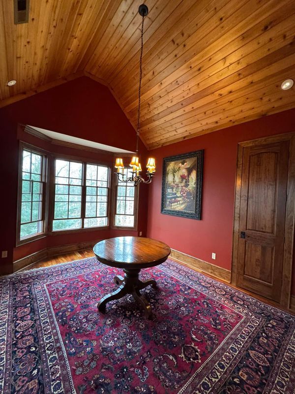 Cozy red-paneled room with vaulted wood ceiling, round table, bay window, chandelier, and patterned rug.