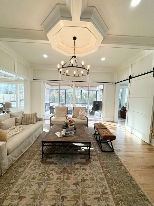 Bright sunroom with white sofas, a coffee table, patterned rug, chandelier, and large windows overlooking outside