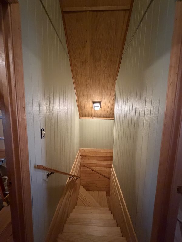 Narrow wooden staircase with a handrail, beige walls, and a ceiling light in a tight hallway.