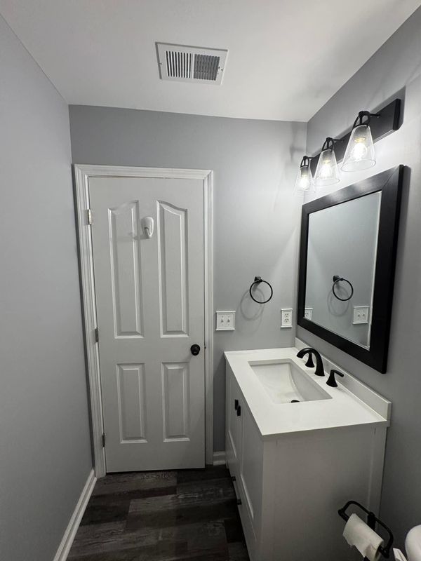 Small gray bathroom with white vanity, black mirror, wall lights, and a closed door.
