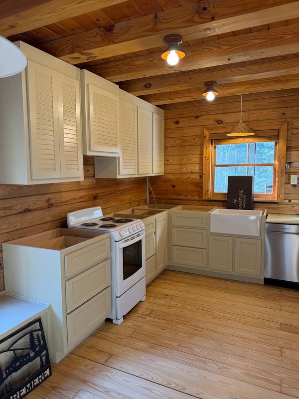 Small rustic kitchen with wood-paneled walls, white cabinets, stove, sink, and a window.