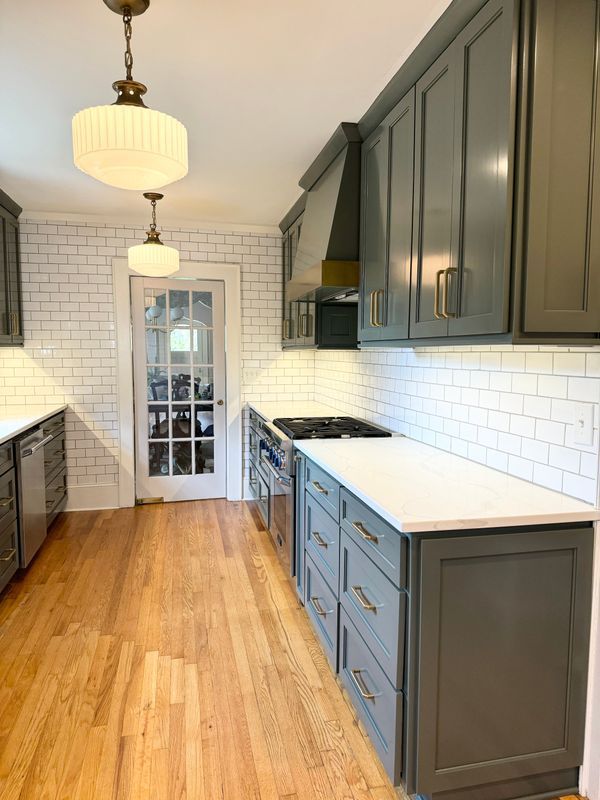 Bright galley kitchen with gray cabinets, white tile backsplash, wood floor, and pendant lights.