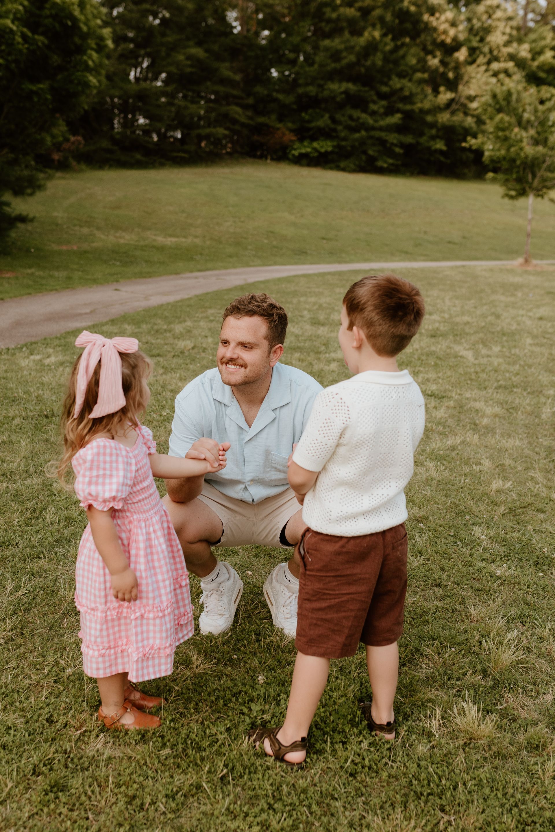 Three people chatting on a grassy park lawn, with a man kneeling and smiling at two children.
