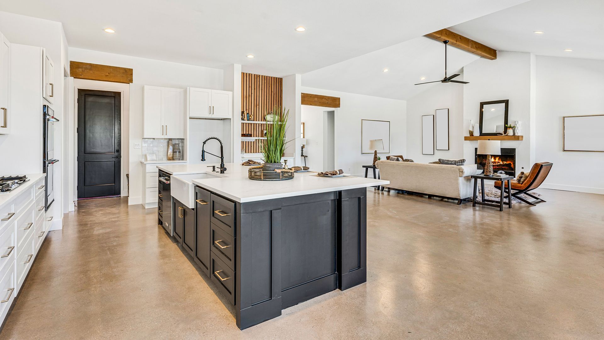 Bright open-plan kitchen and living room with white walls, dark island, wood beams, and minimalist decor.