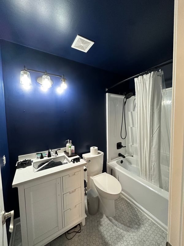Navy blue bathroom with white vanity, toilet, bathtub shower, and bright vanity lights.
