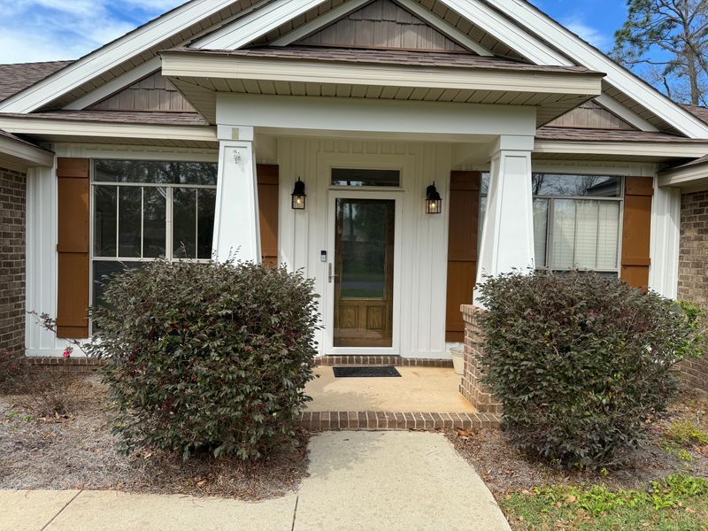 Front entrance of a suburban house with white columns, brown shutters, and trimmed bushes.