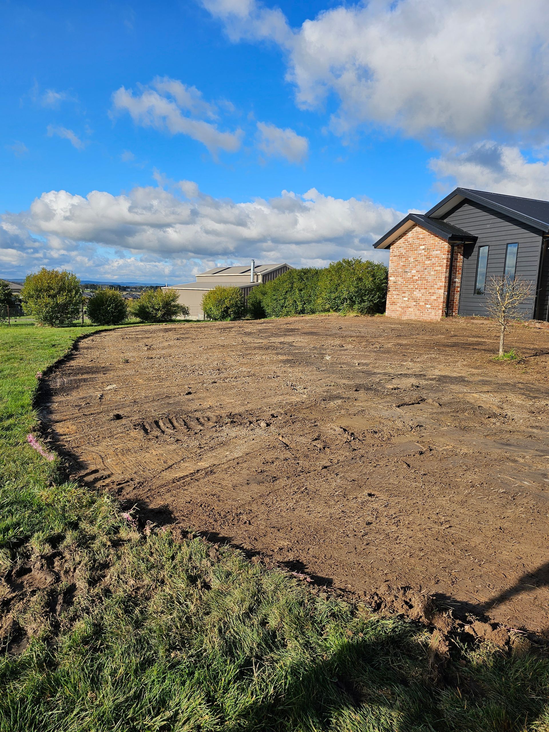 A newly tilled garden bed with a curved border sits beside a green lawn, a brick house, and a blue sky with clouds.