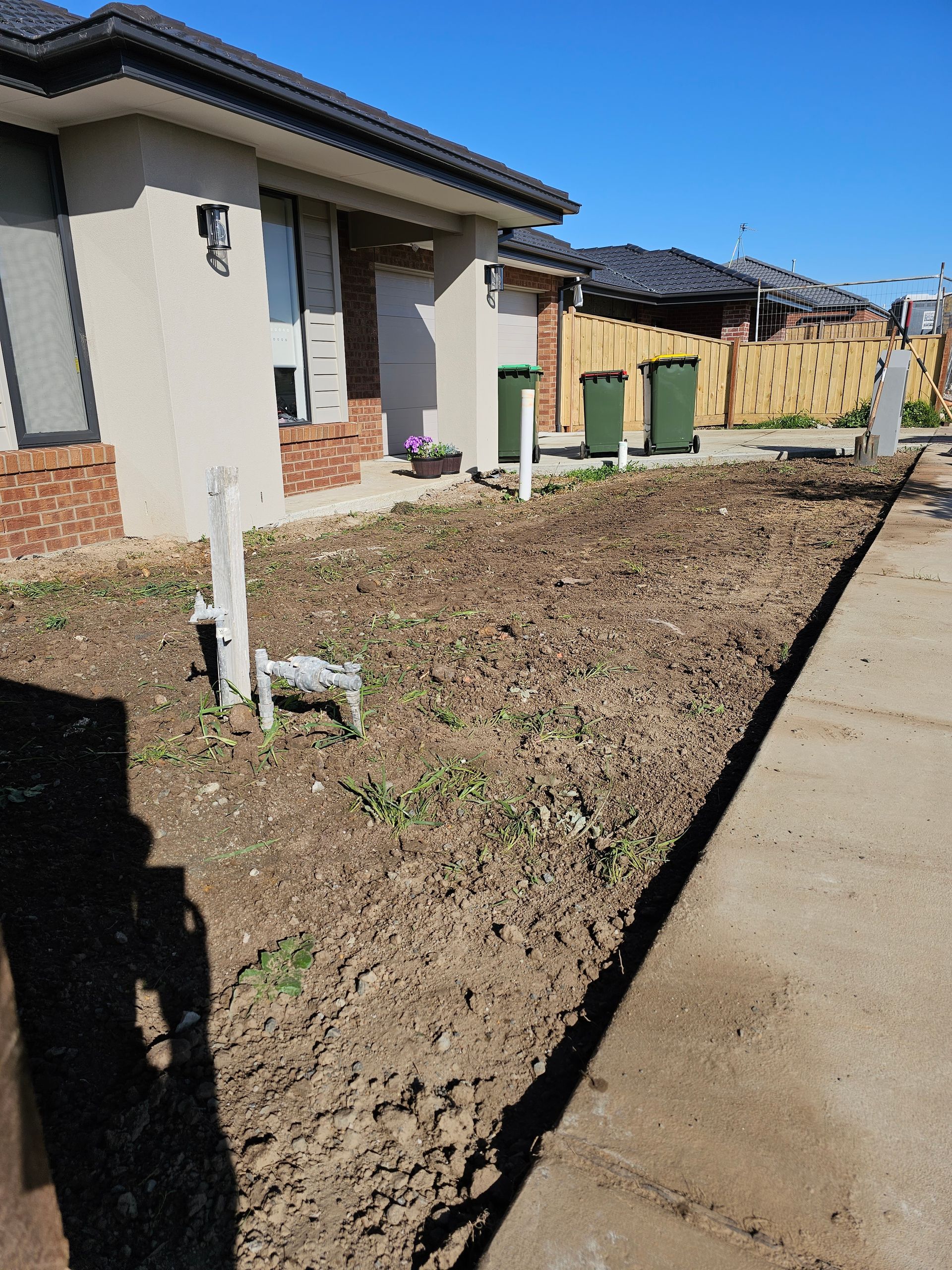 Exterior view of a house with a bare dirt front yard, green trash bins, and a concrete sidewalk under a blue sky.