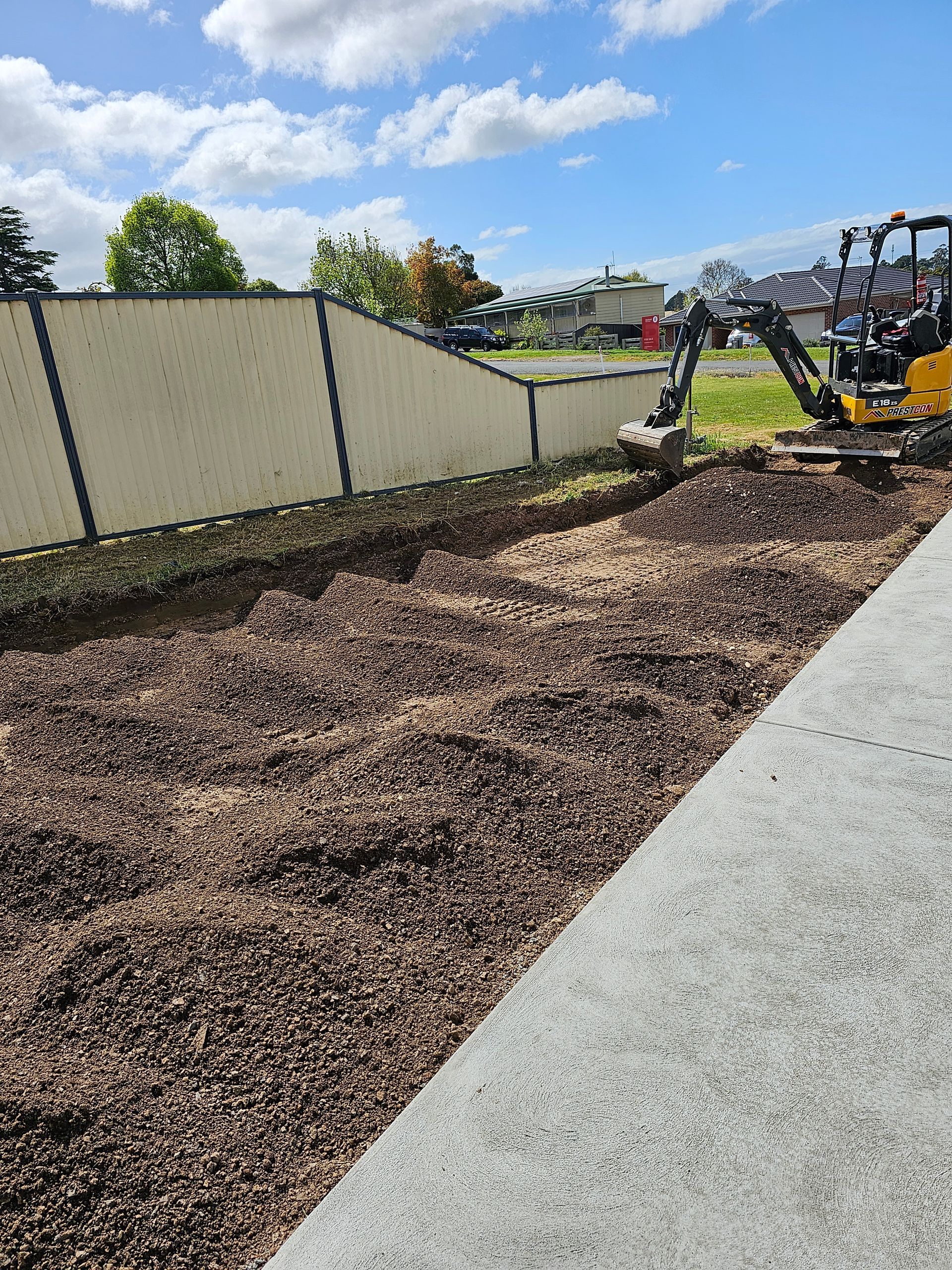 A small excavator digs in a long, gravel-filled trench alongside a concrete pathway under a partly cloudy sky.