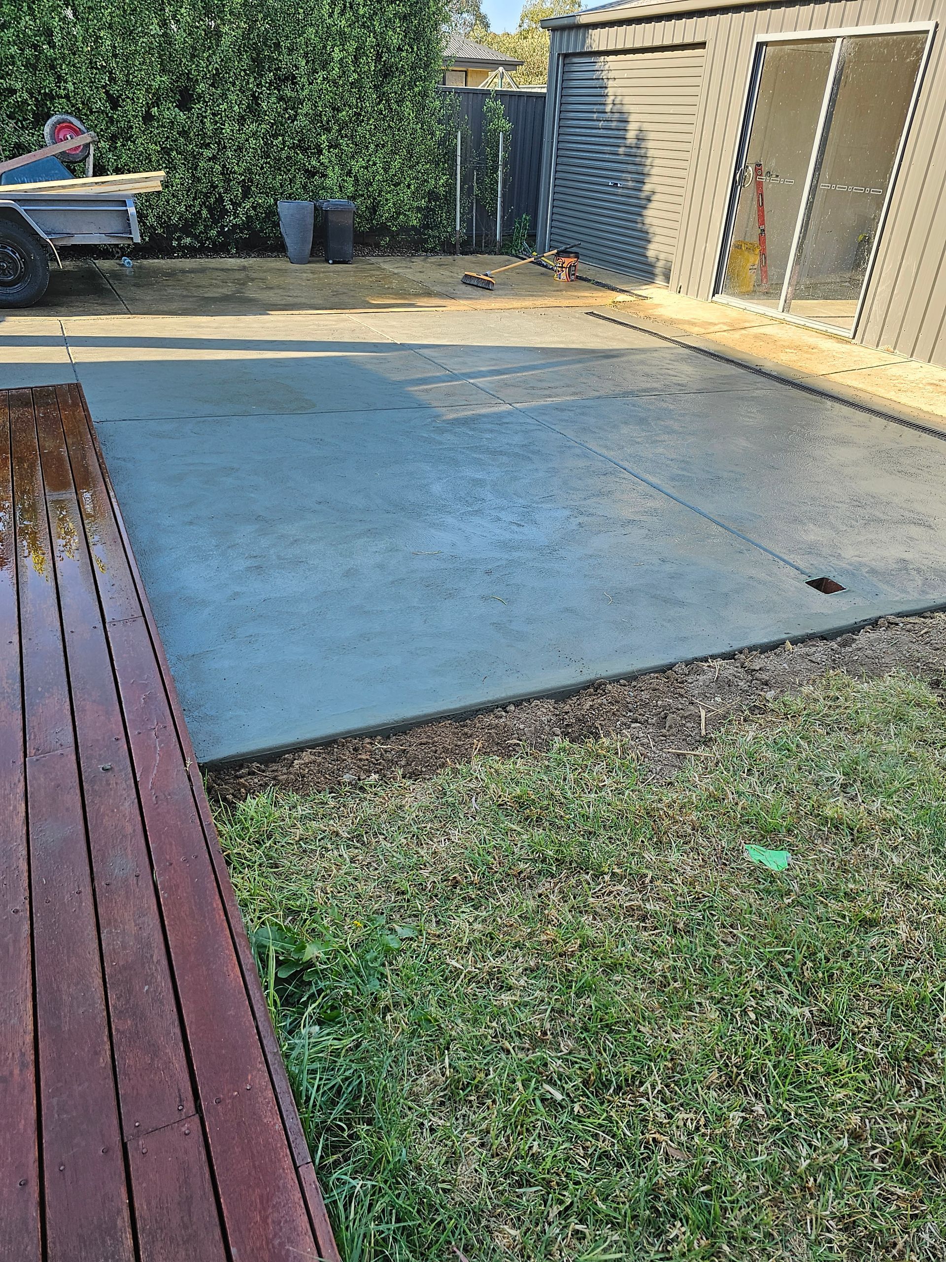 A concrete patio bordered by grass, with a wooden deck at the left. Shed in the background.