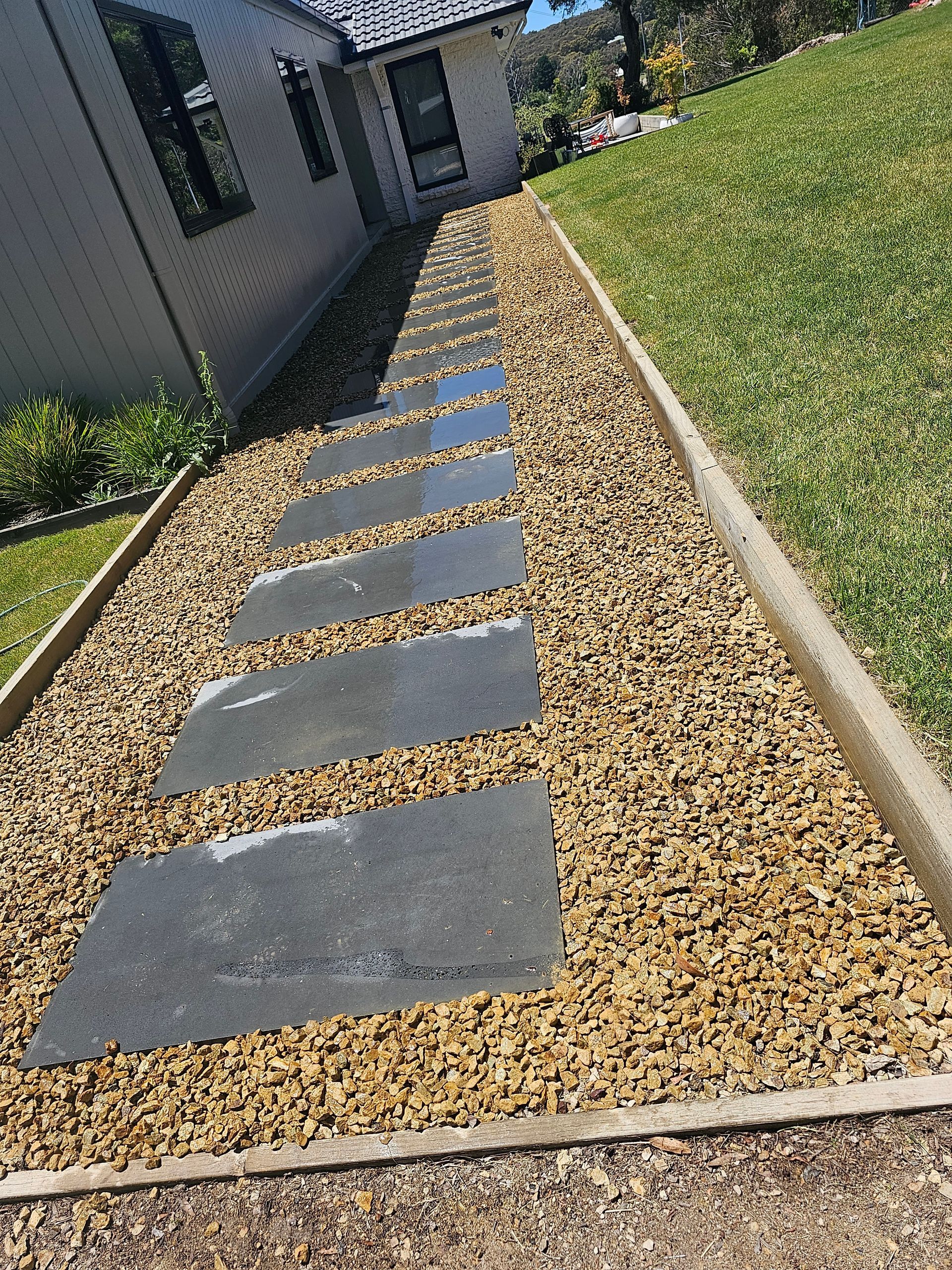 Slate stepping stones bordered by tan gravel and wood planks lead to a house, with green grass lining the path.
