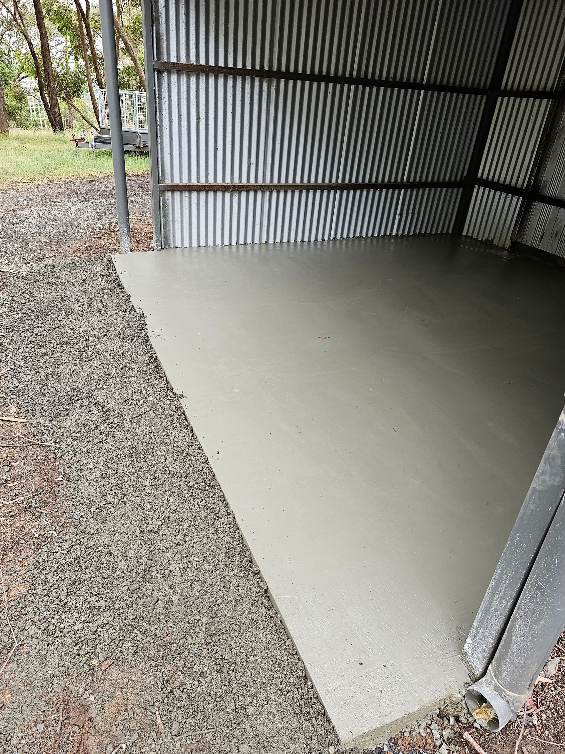 A freshly poured concrete floor inside a corrugated metal shed. Gravel borders the concrete on the left.