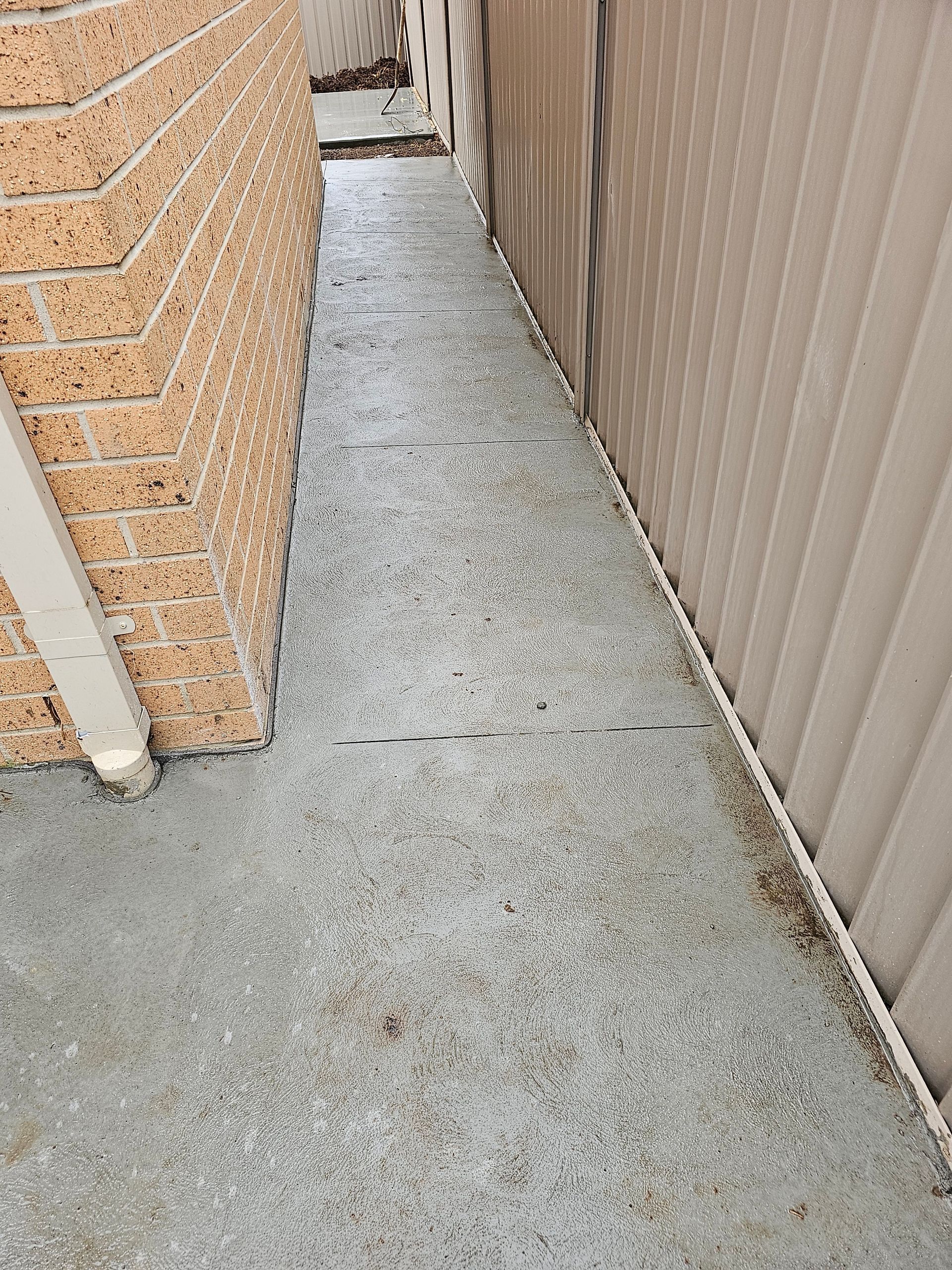 Narrow concrete pathway between a brick wall and a beige corrugated fence. Small debris is scattered on the surface.