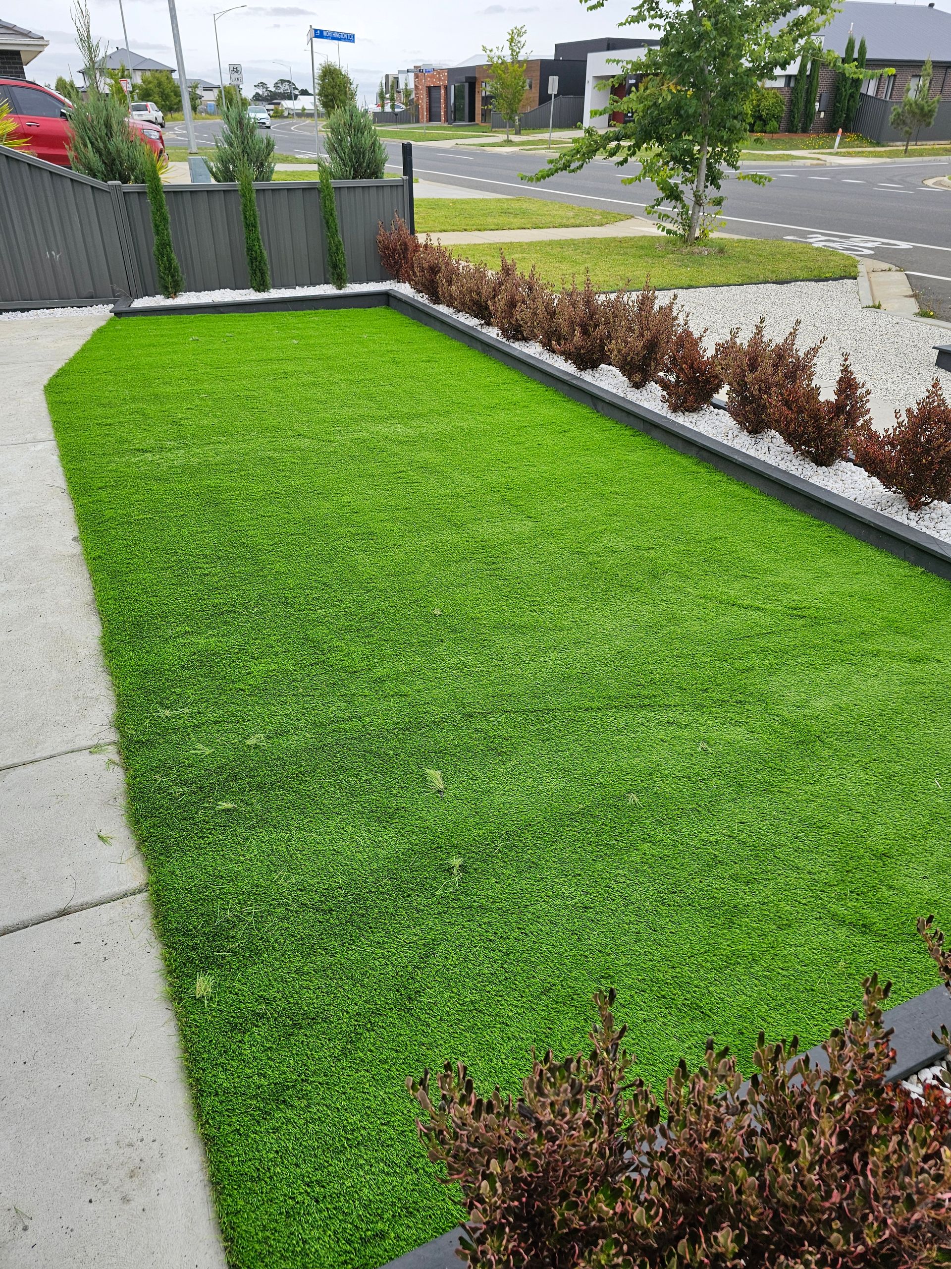 Lush green artificial lawn bordered by dark gray edging, white stones, and red bushes, in front of a gray fence and sidewalk.
