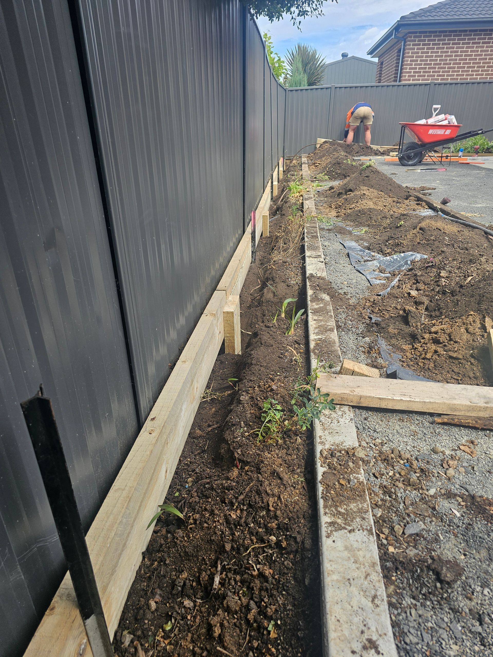 A person working in a backyard, digging a trench next to a dark fence. Wooden beams border the trench.