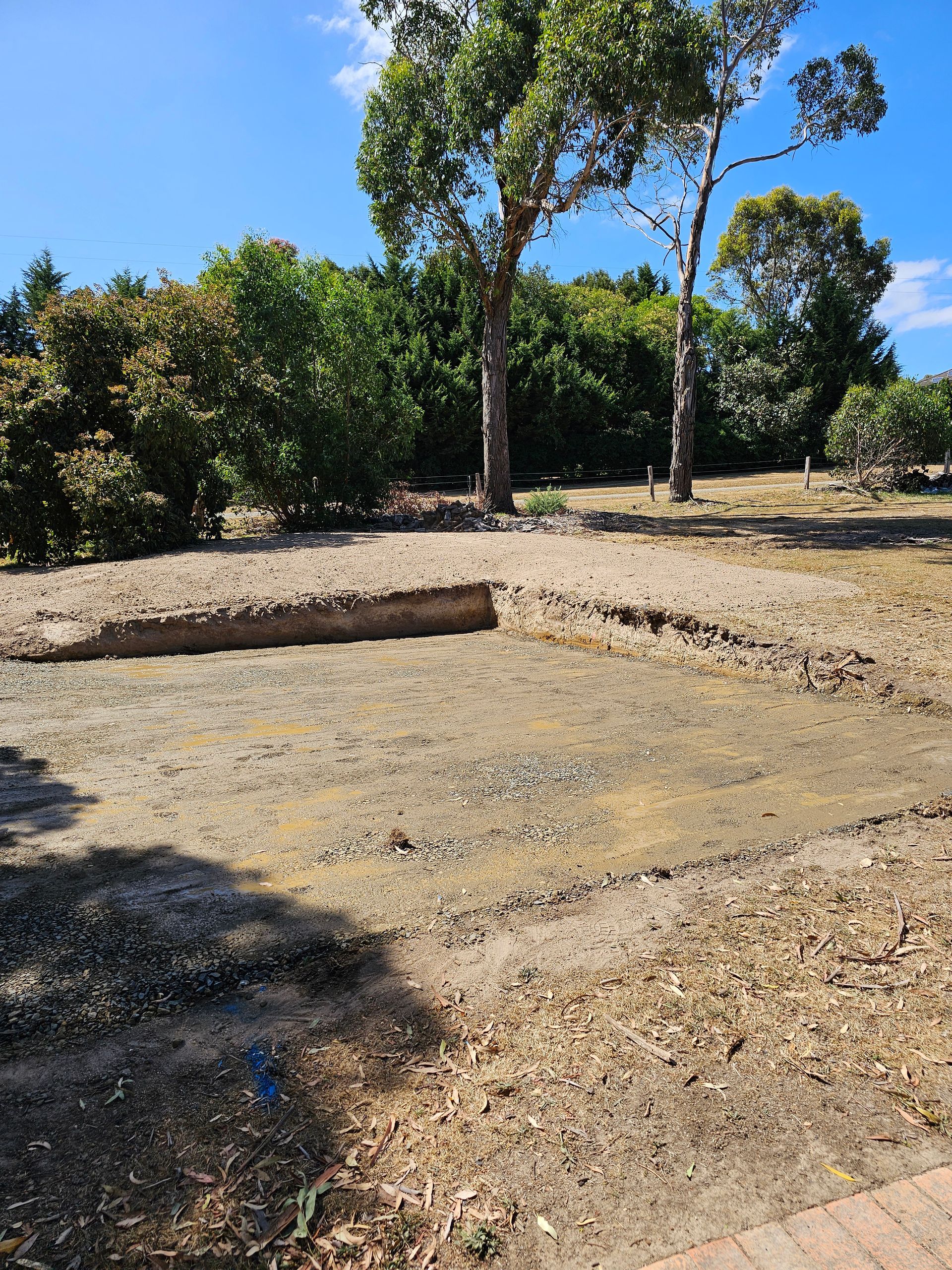 A rectangular excavation in a yard with surrounding trees and a blue sky. Dirt and gravel fill the hole.