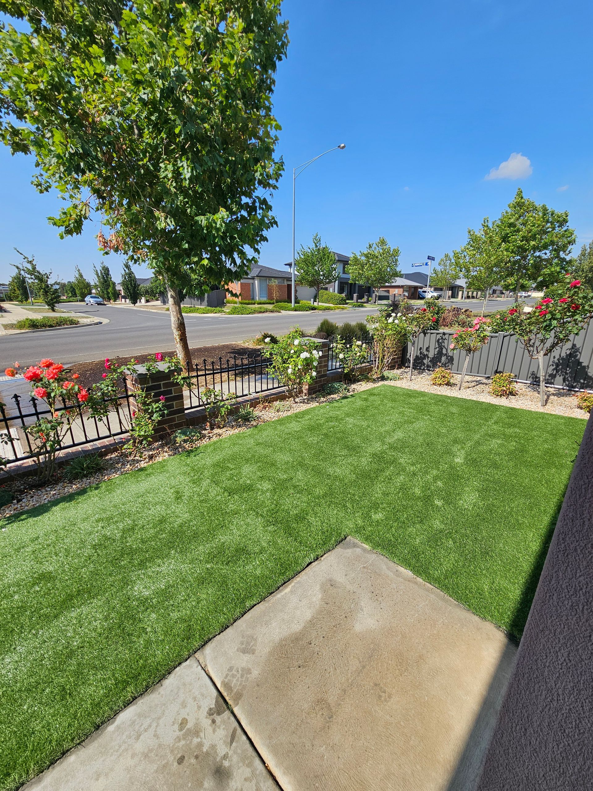A small, grassy front yard with colorful flower beds and a sidewalk, under a bright blue sky. A tree provides shade.