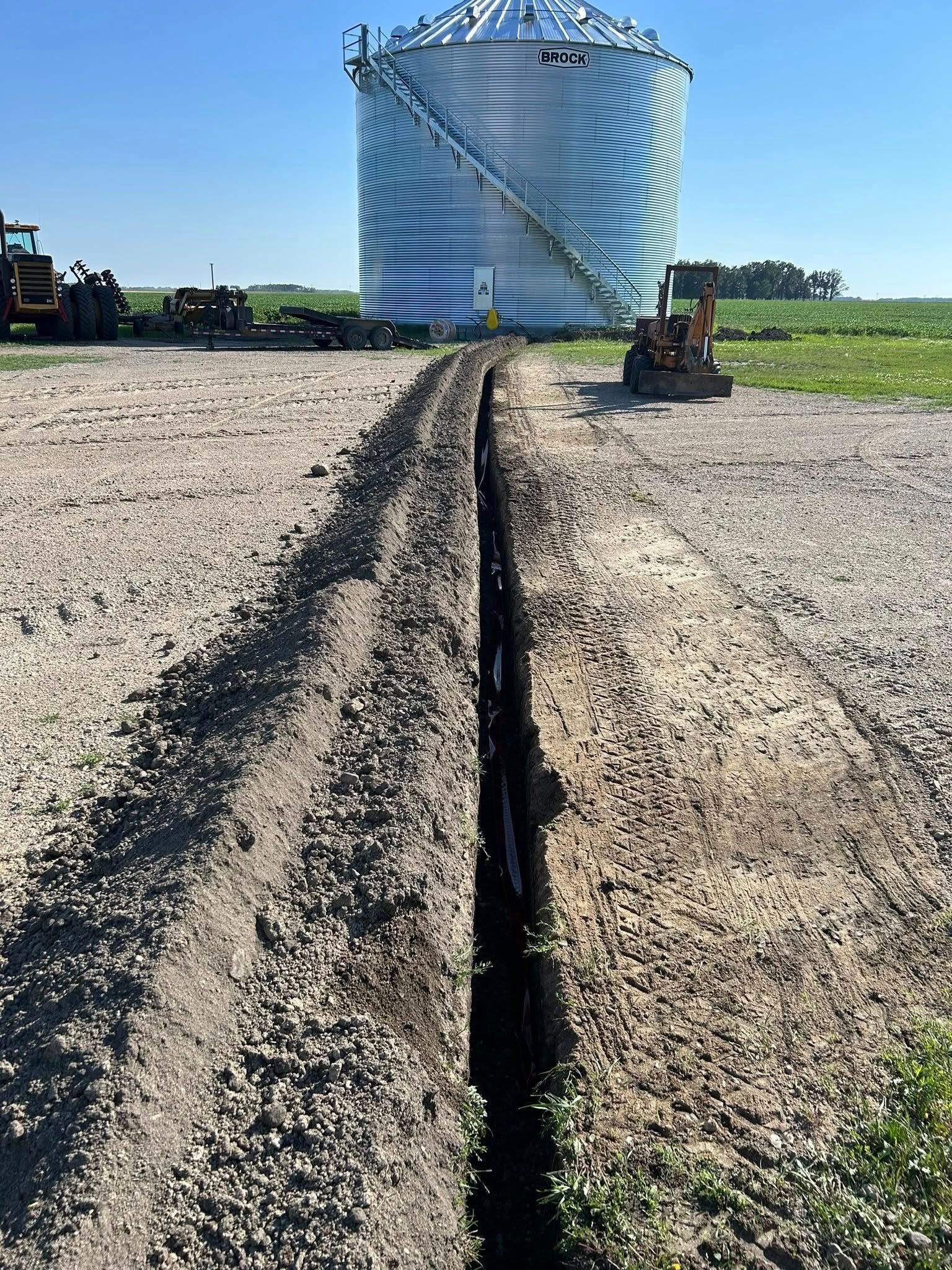 A trench dug in gravel leads to a large silver grain silo. Machinery sits in the background on a sunny day.