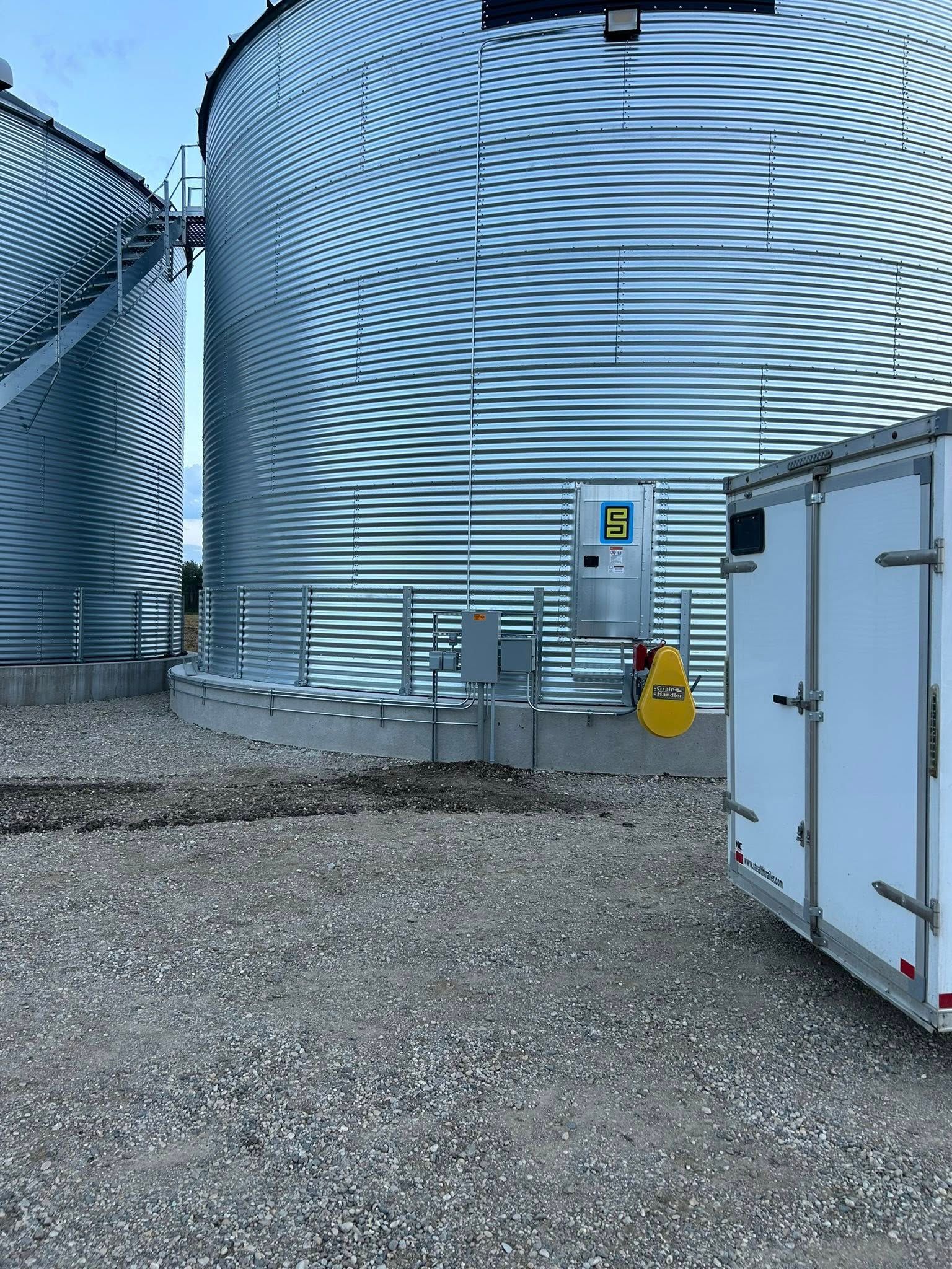 Two large grain silos next to a white trailer. Control box and gravel ground.