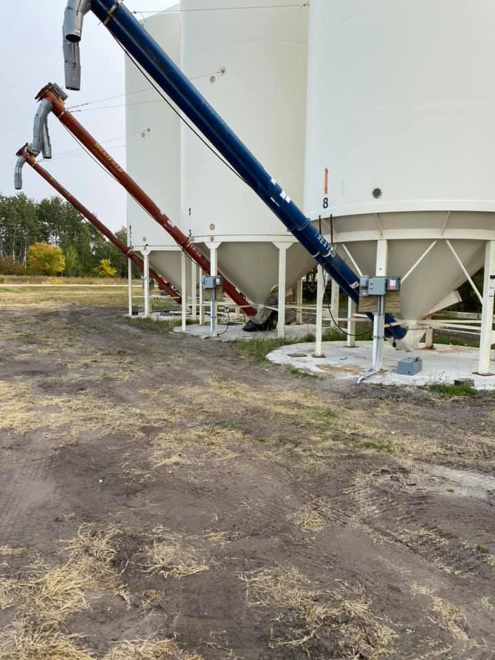 Three white grain silos with blue and red augers against a field and trees.