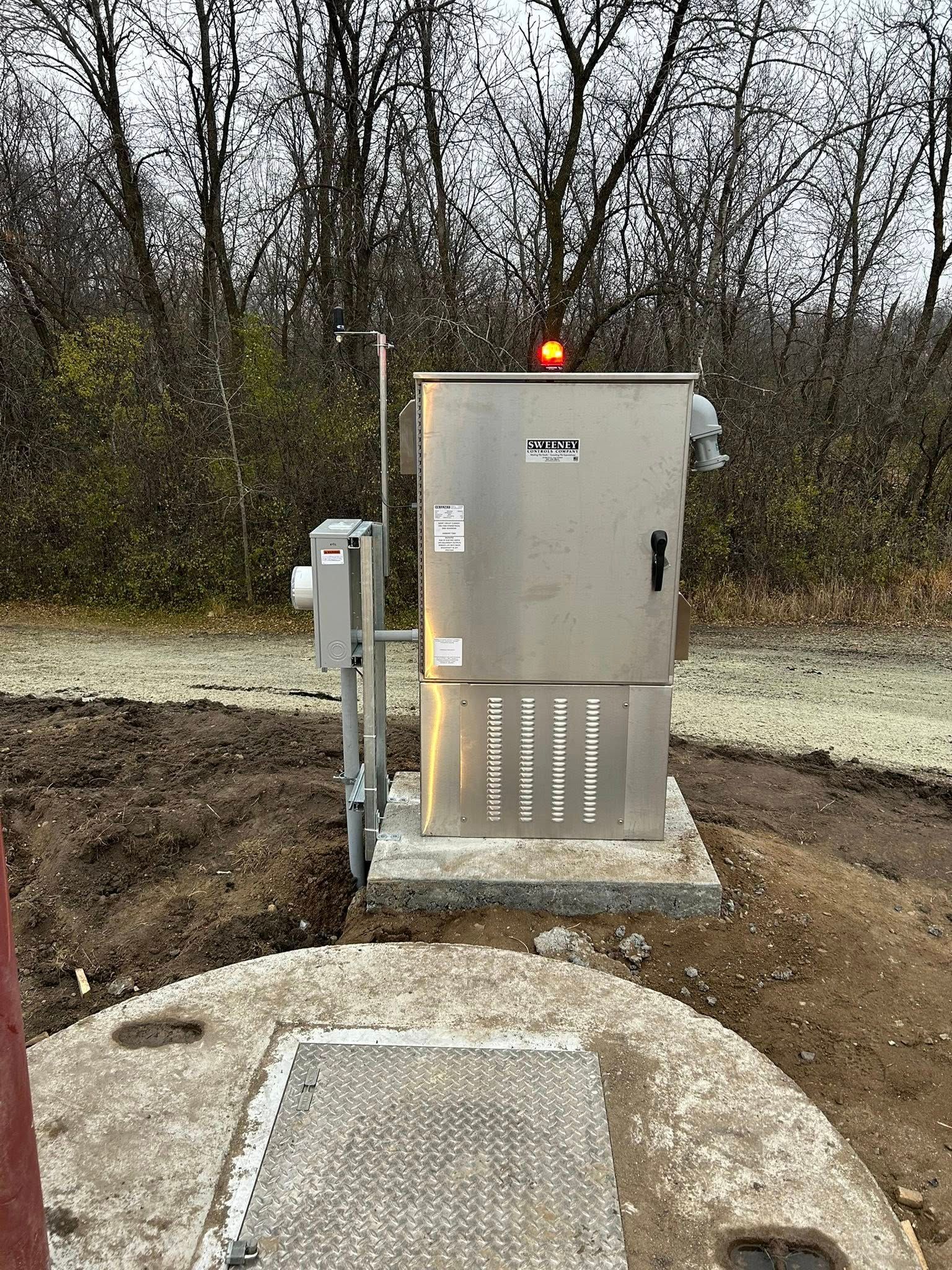 A stainless steel electrical box with an orange light on a concrete pad next to a manhole cover in a wooded area.