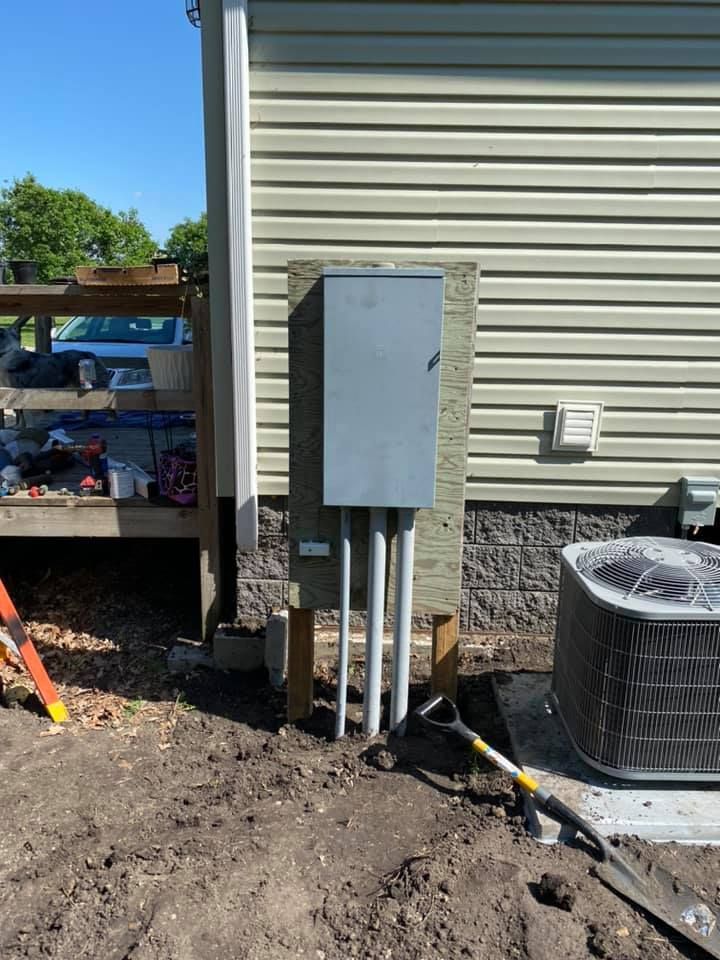 Electrical panel mounted on wood siding, with conduit pipes extending into the ground.
