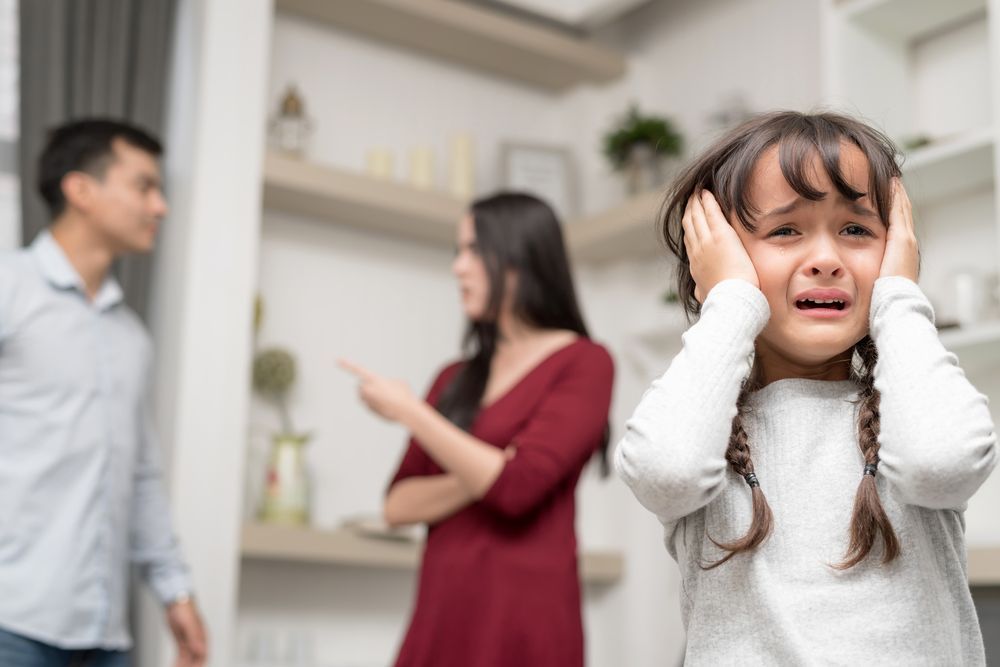 A little girl is crying while her parents argue in the background.