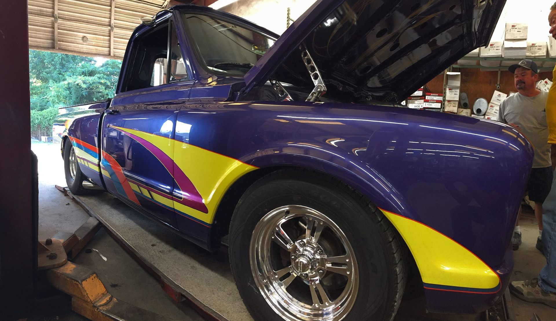 A purple and yellow truck with the hood up is parked in a garage.