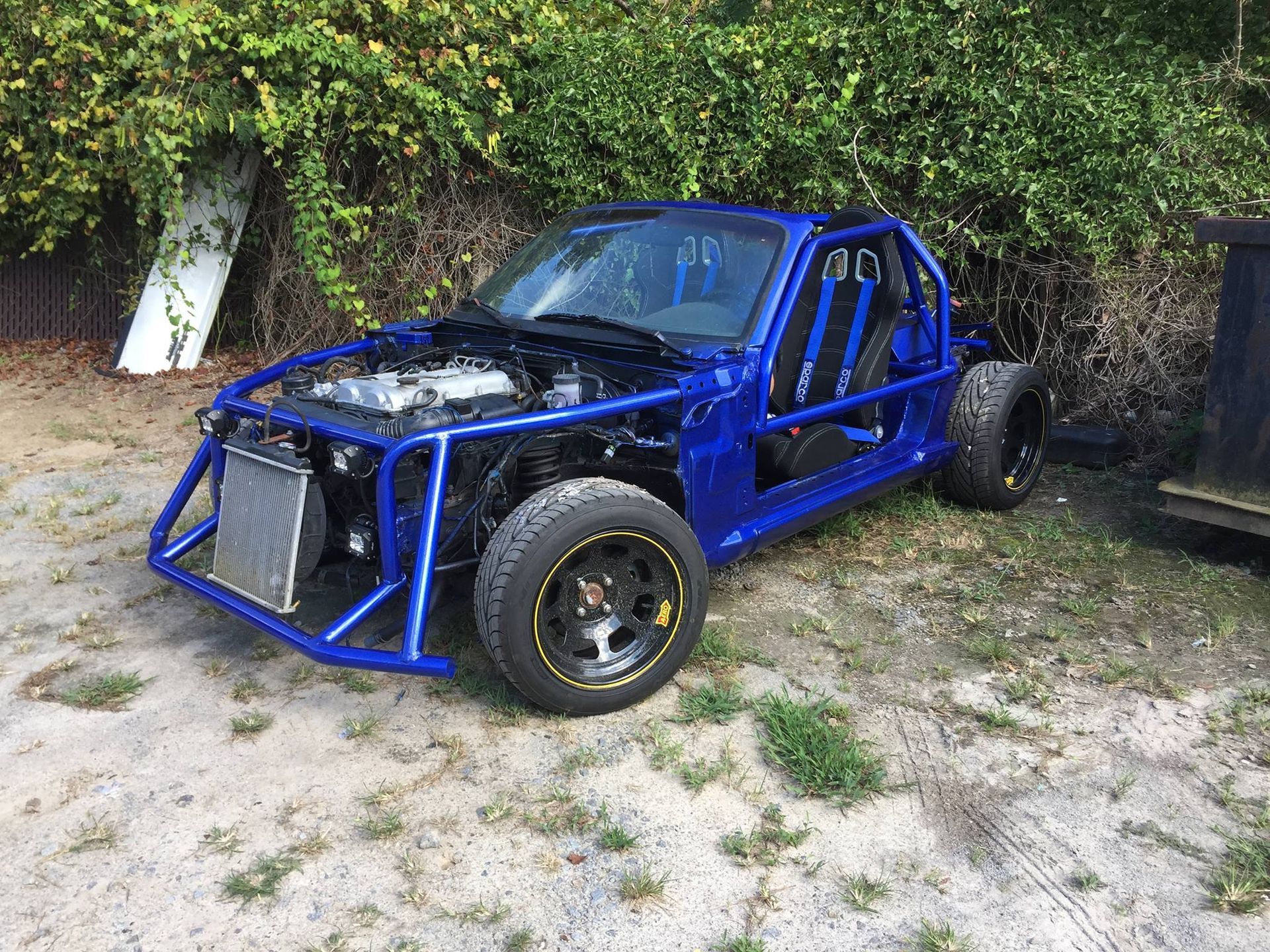 A blue car with the hood open is parked in a dirt field.