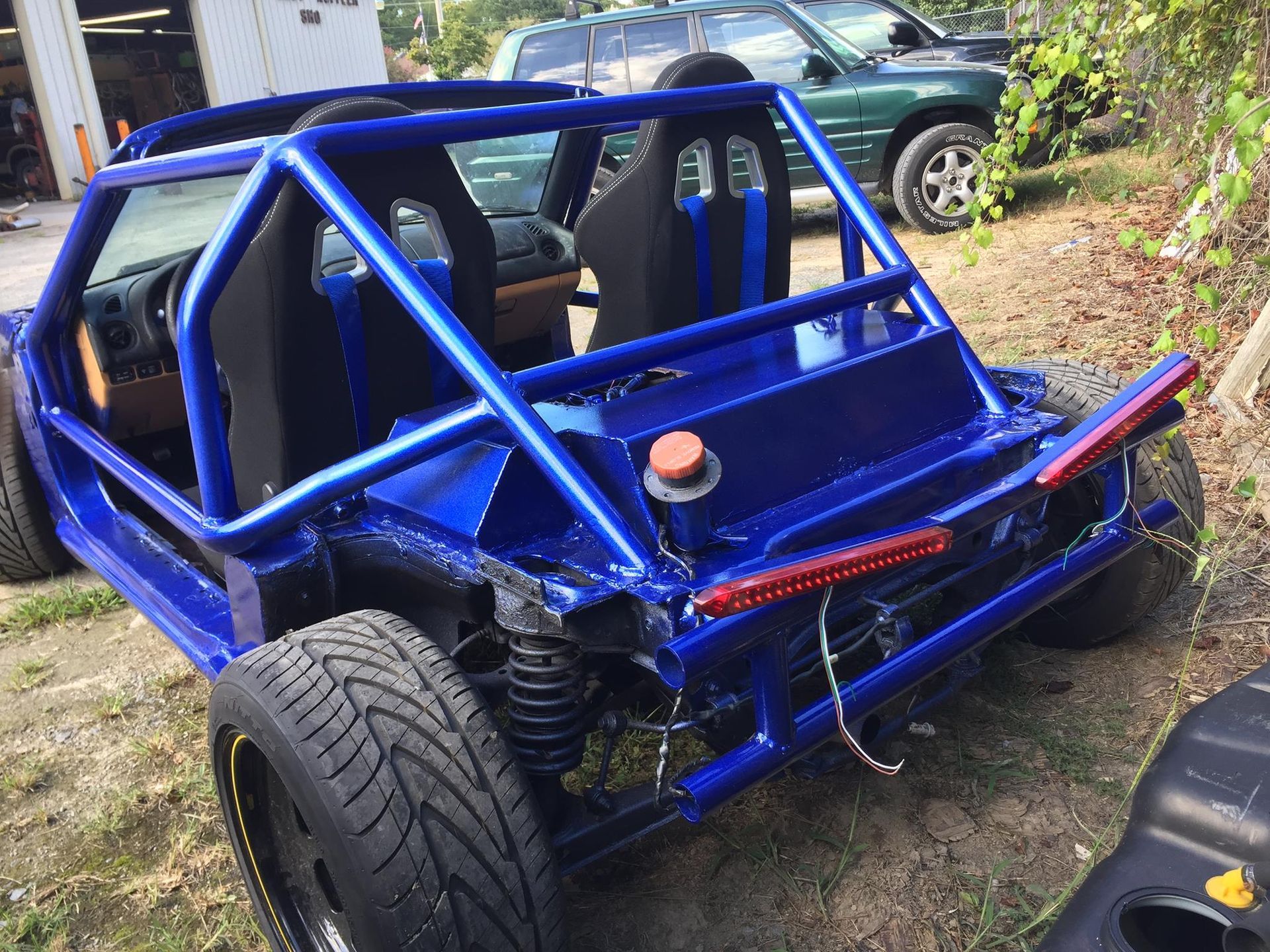 A blue dune buggy is parked in the dirt in front of a garage.