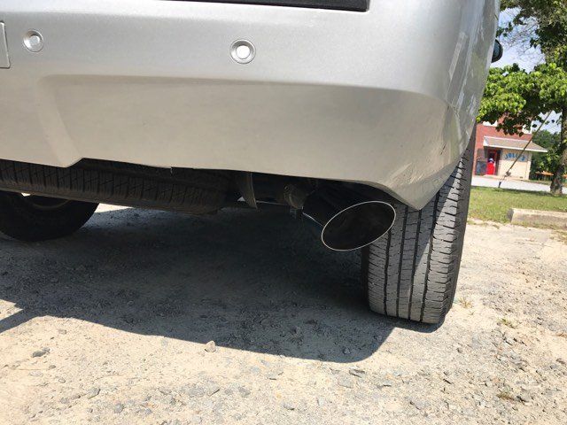 A silver car with a black exhaust pipe is parked on a dirt road.