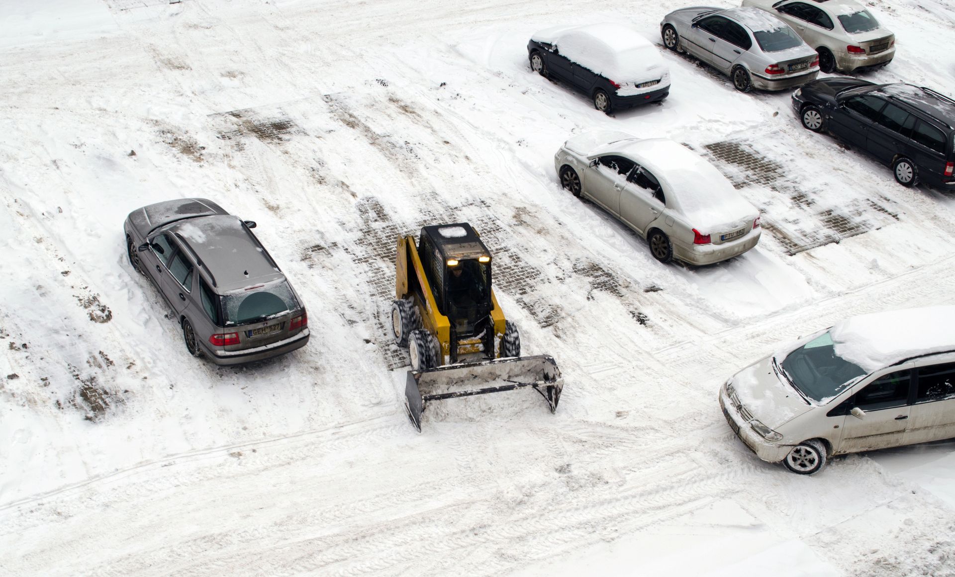 A yellow skid-steer loader clears snow in a parking lot between rows of snow-covered cars.