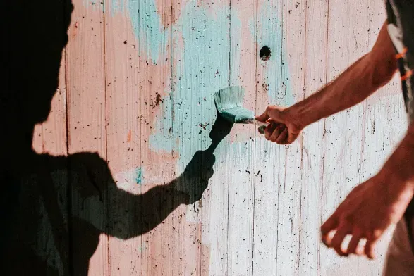 Person painting a wooden wall with a turquoise brush, casting a shadow.