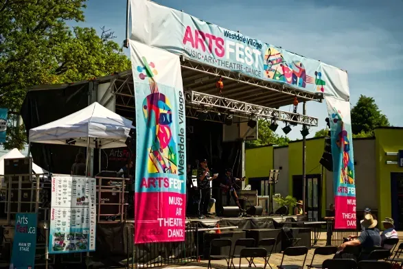 Outdoor Arts Fest stage with band playing; banners, signage, people in audience, sunny.