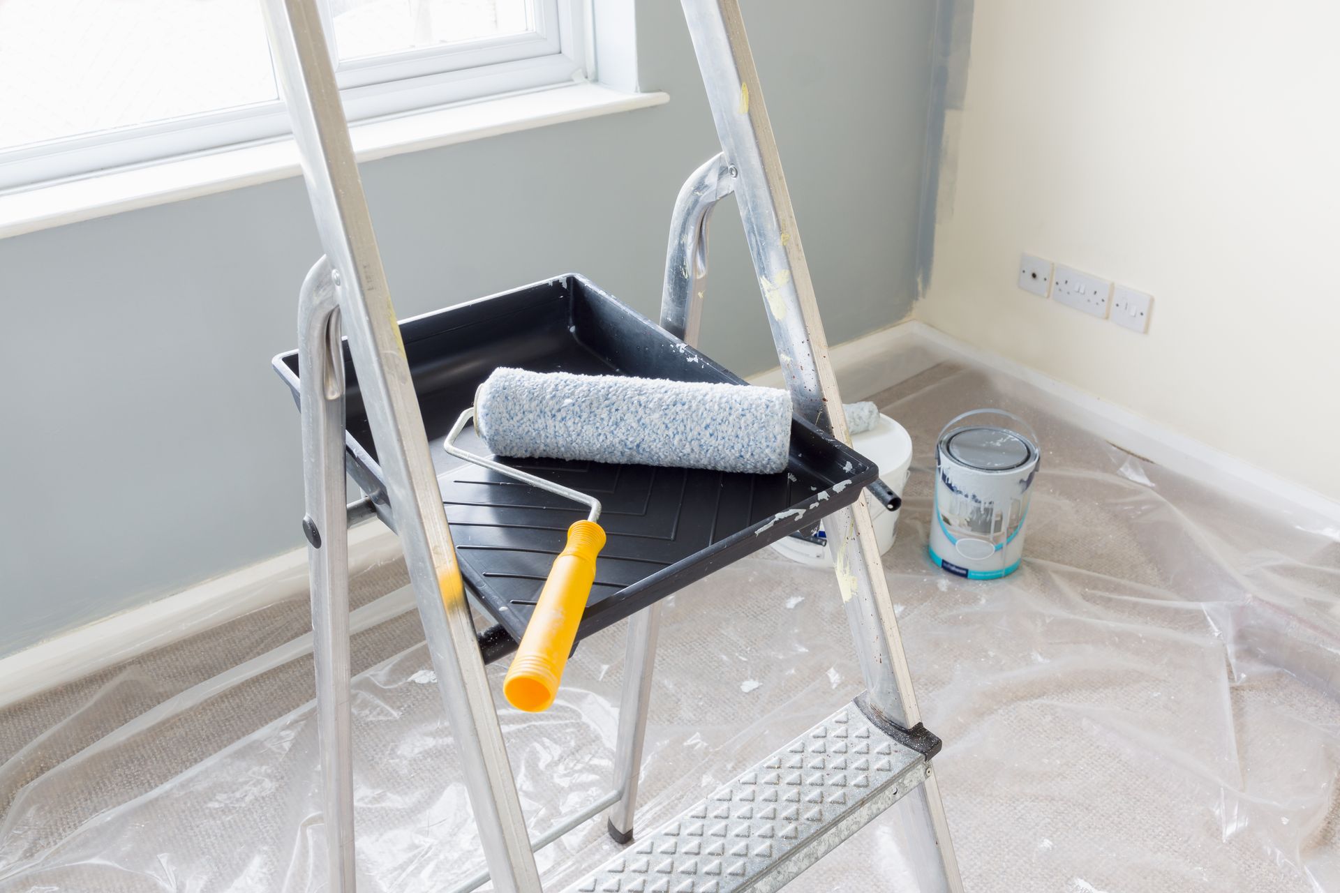 Paint tray and roller on a step ladder in a room being painted, with plastic floor covering and a paint can nearby.