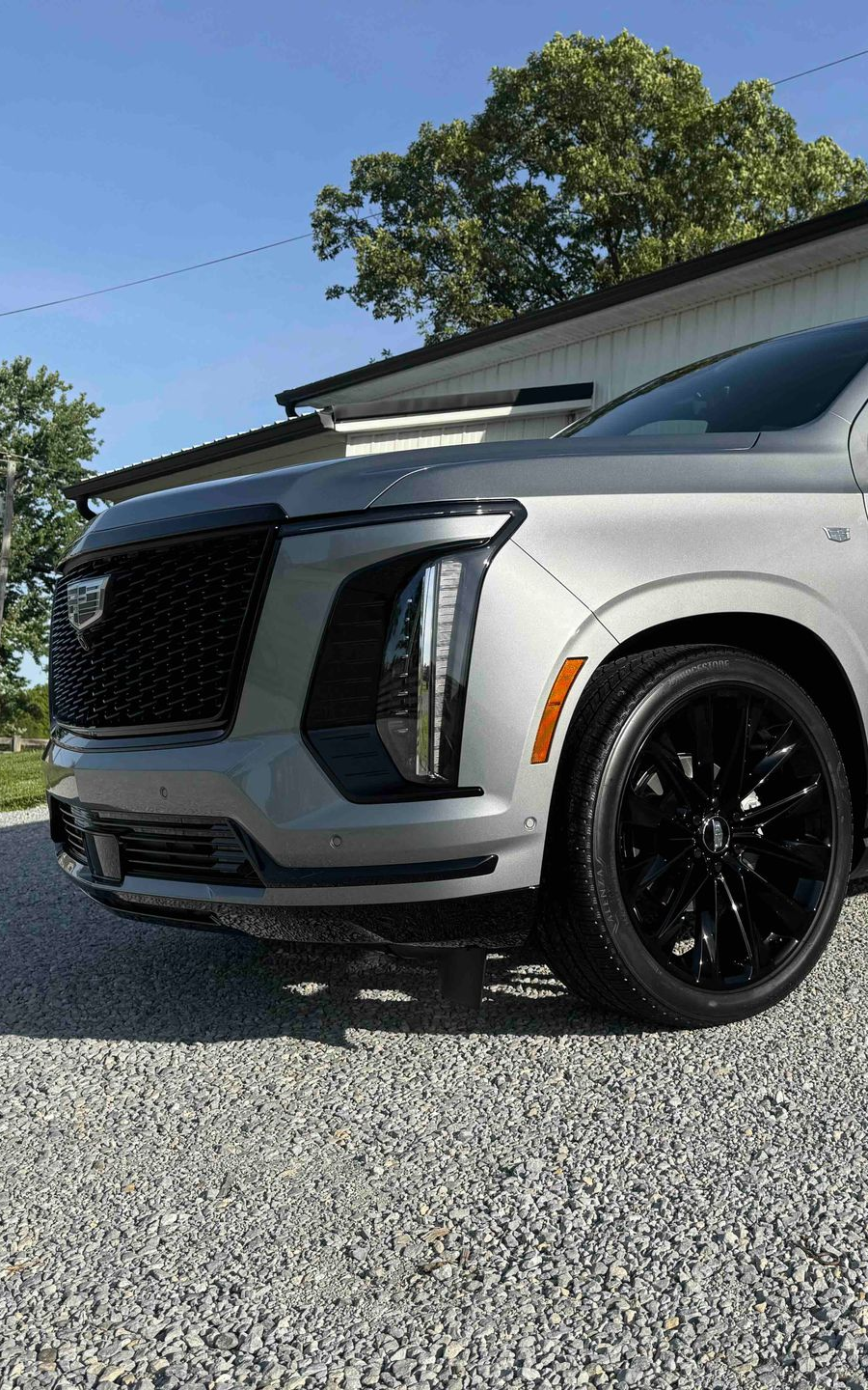 Silver Cadillac Escalade with black wheels parked on gravel, against a building on a sunny day.
