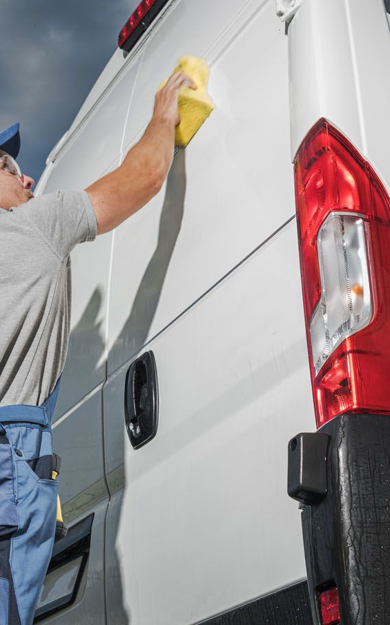 Man cleaning a white van with a yellow sponge. Red taillight and dark sky in background.