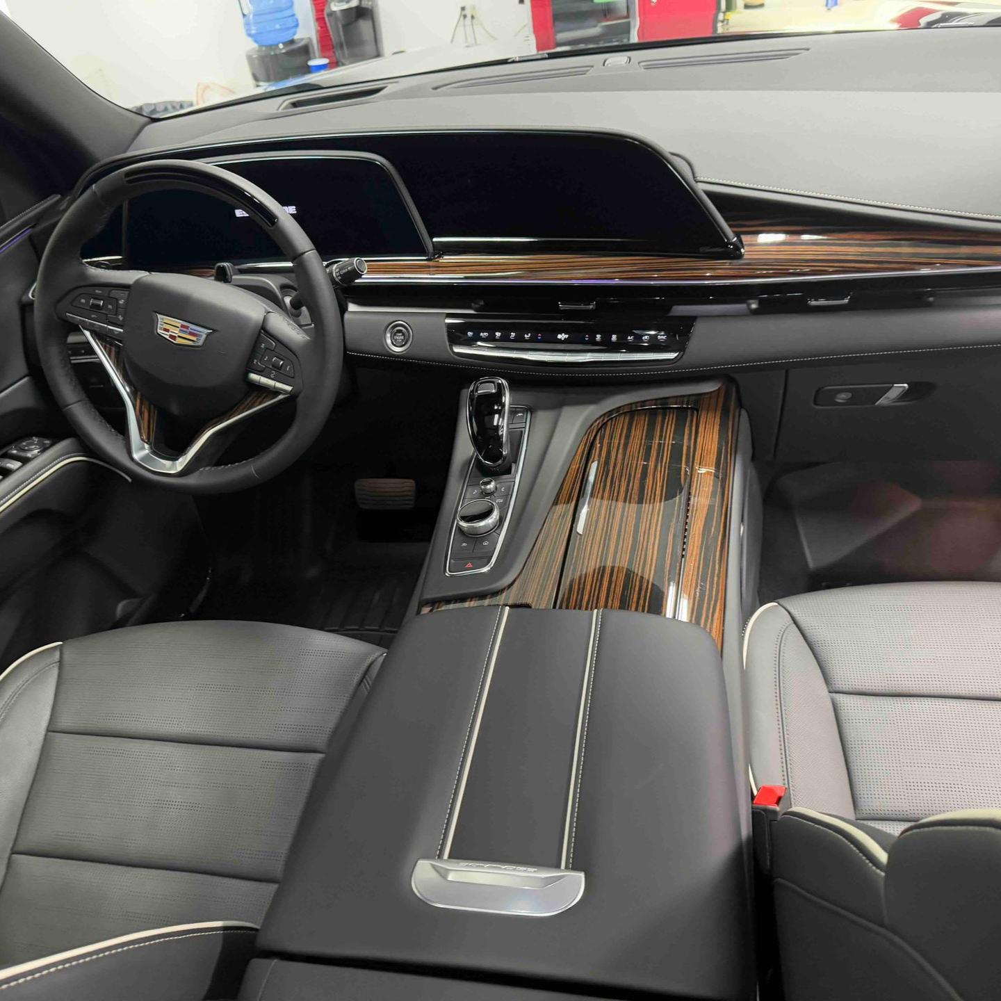 Interior view of a Cadillac SUV; black leather seats, wood trim, large infotainment screen, steering wheel, and center console.