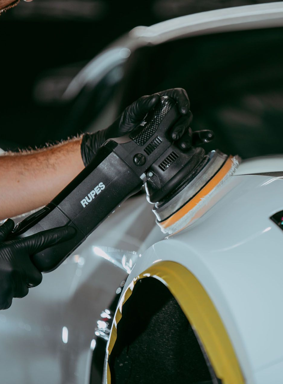 Person polishing a car with a rotary buffer, hands on the machine, black car reflects the process.