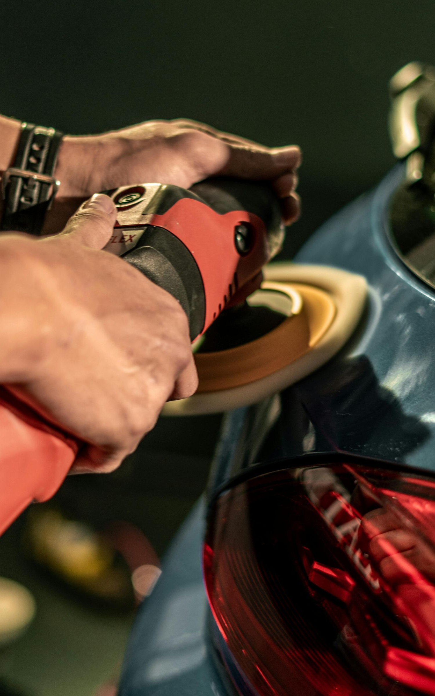 Person using a red and black power buffer on a blue car's tail light.