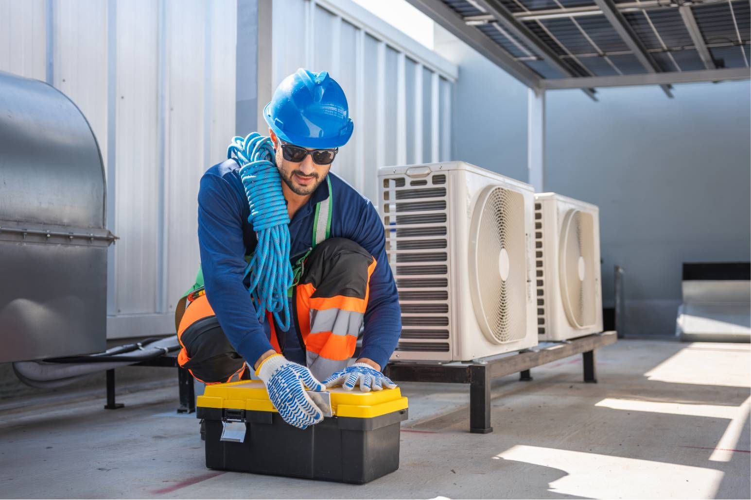 Worker in blue hard hat kneeling by rooftop HVAC units, checking a toolbox on a sunny industrial roof