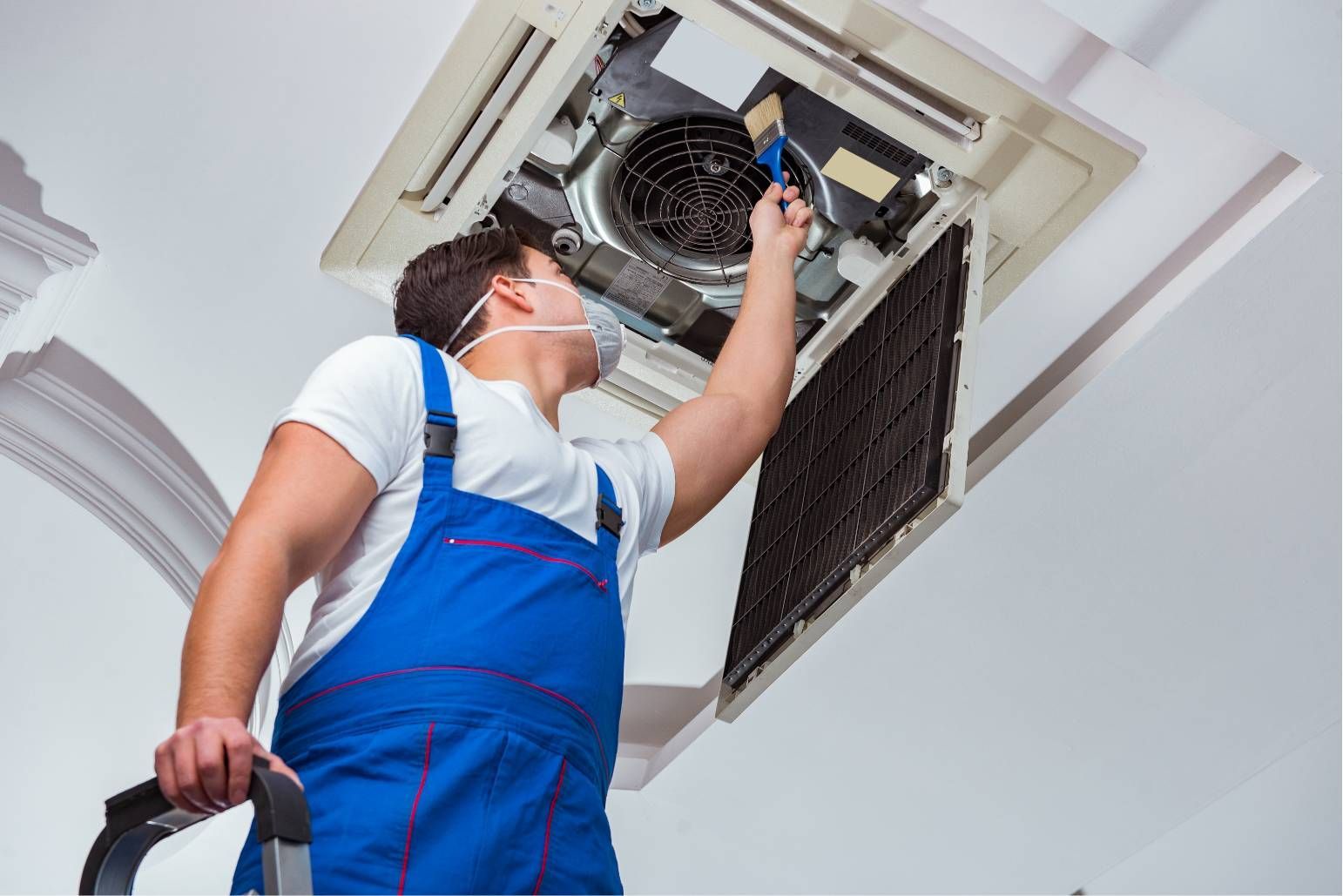 Technician in blue overalls repairing a ceiling air conditioner with a screwdriver