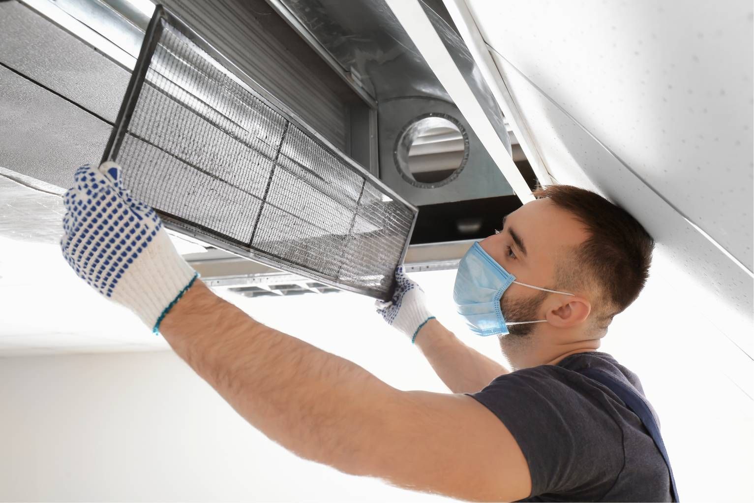 Masked worker cleaning a ceiling air vent with a cloth and gloved hand