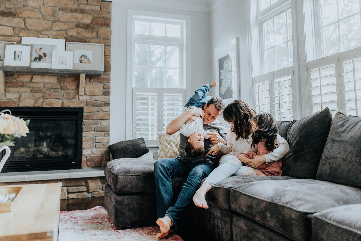Two children on a gray couch with a dog in a bright living room near a stone fireplace